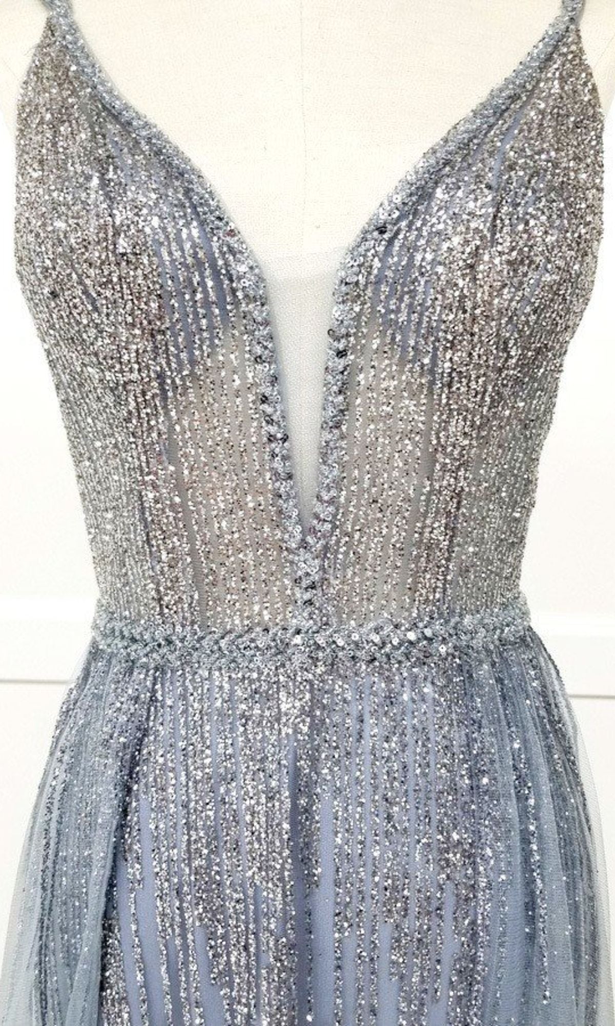A close-up of the Sheer-Waist Long Sequin Prom Dress (Chicas YG5021), a sleeveless silver evening gown with a deep V neckline, sheer mesh panel, and glittering sequin print, displayed on a dress form.